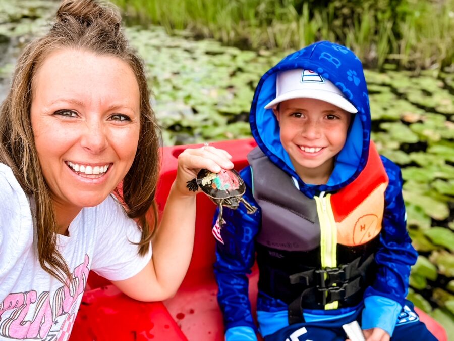 A mother holding a turtle next to a son smiling in a paddle-boat on the pond.